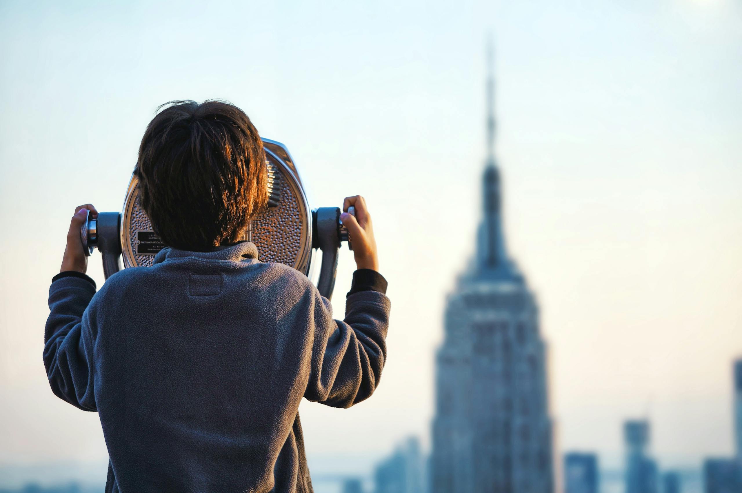 A child uses binoculars to view the Empire State Building in New York City at sunset.