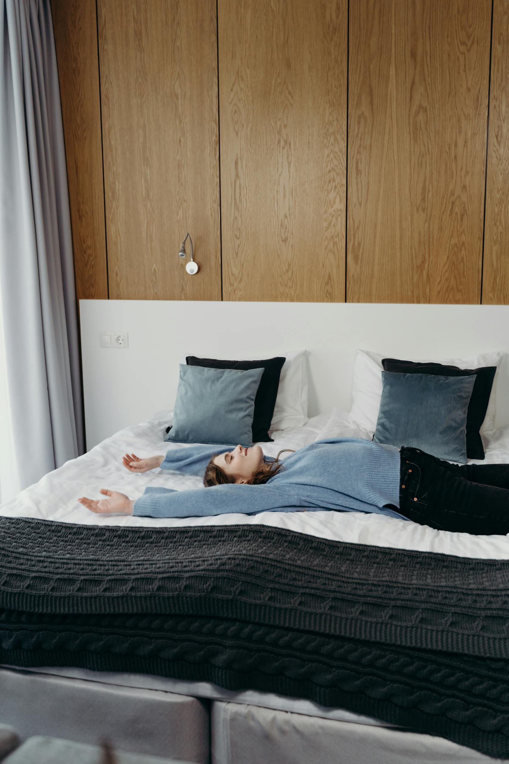 A woman in casual attire lies relaxed on a bed in a stylish, modern bedroom with wooden wall panels.