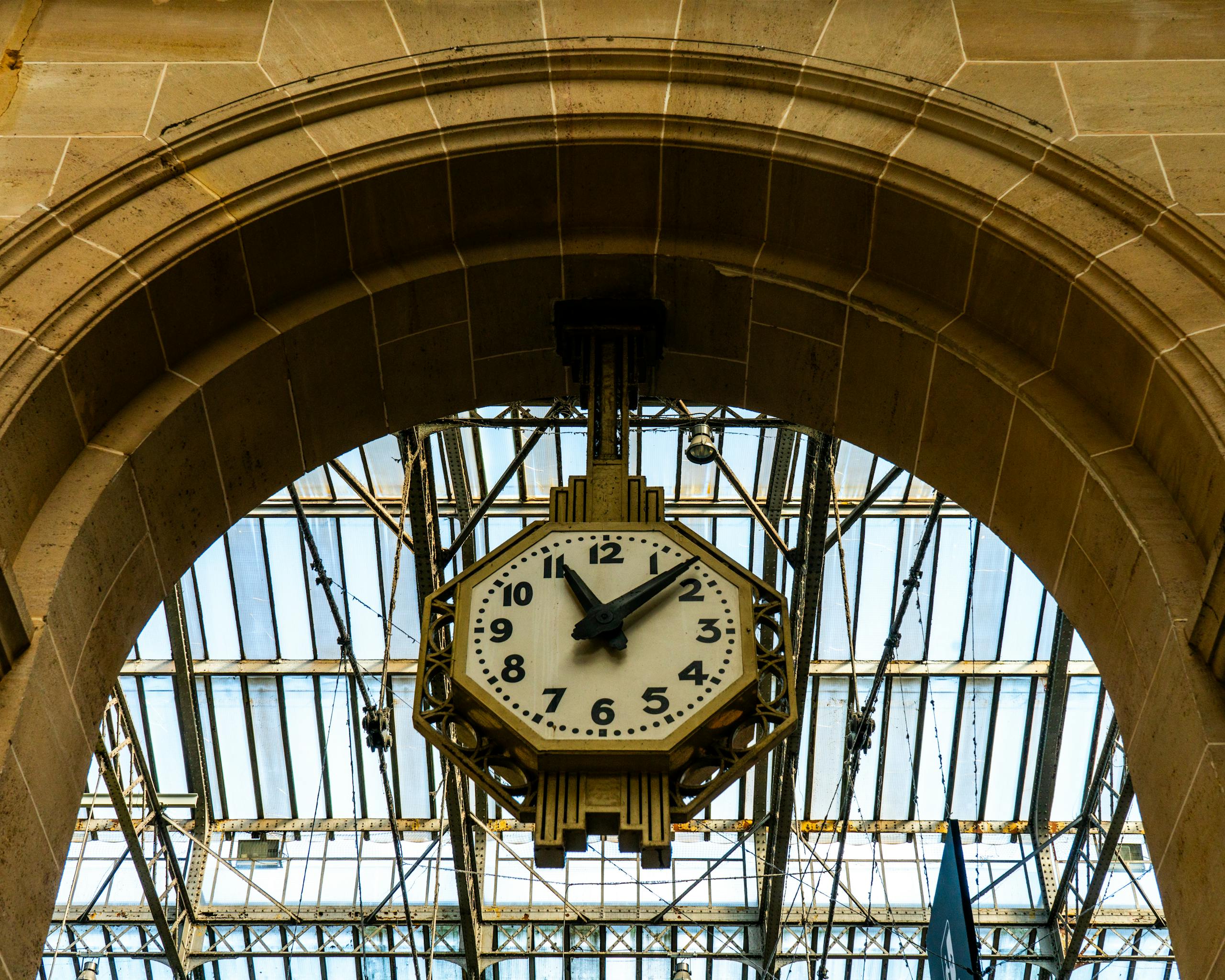 Industrial-style clock beneath an arch with glass ceiling in a train station setting.